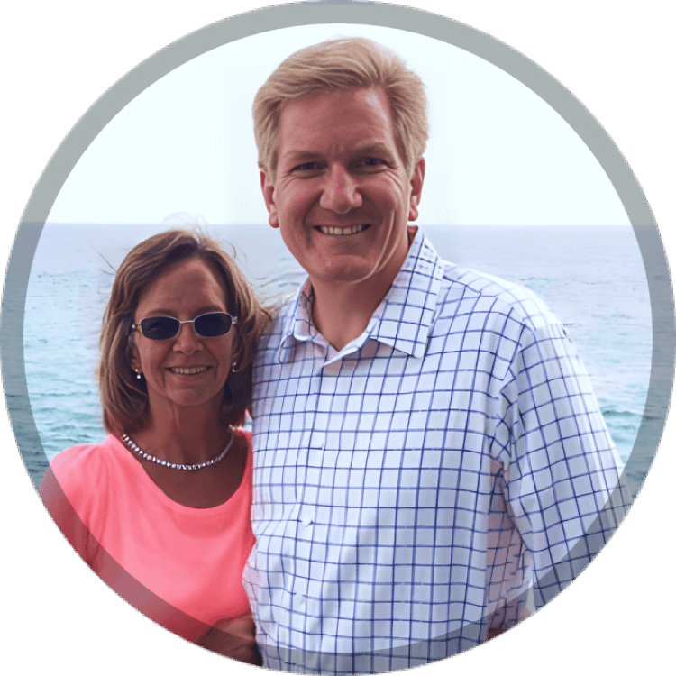 Two smiling adults posing together outdoors with ocean water visible in background.