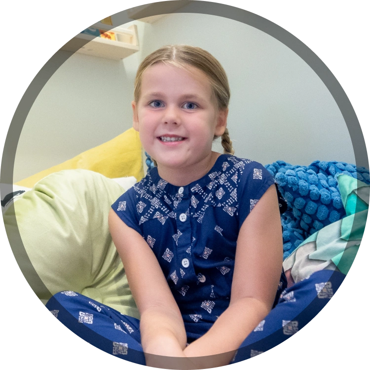 Young girl smiling at camera, sitting on blue patterned couch indoors.